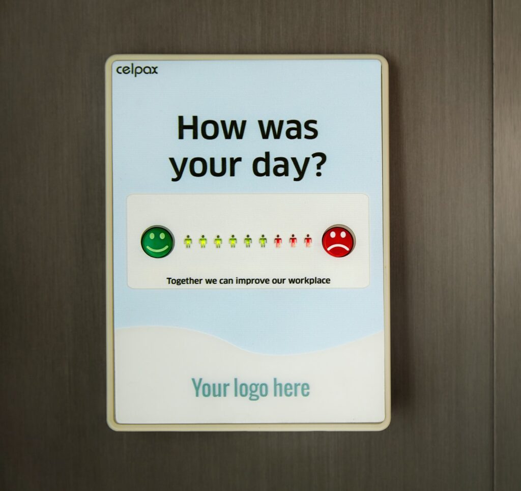 Feedback board on the wall of a workplace that asks employees "How was your day?" and lets them tap a green happy face through to a red sad face to gauge and reduce staff turnover